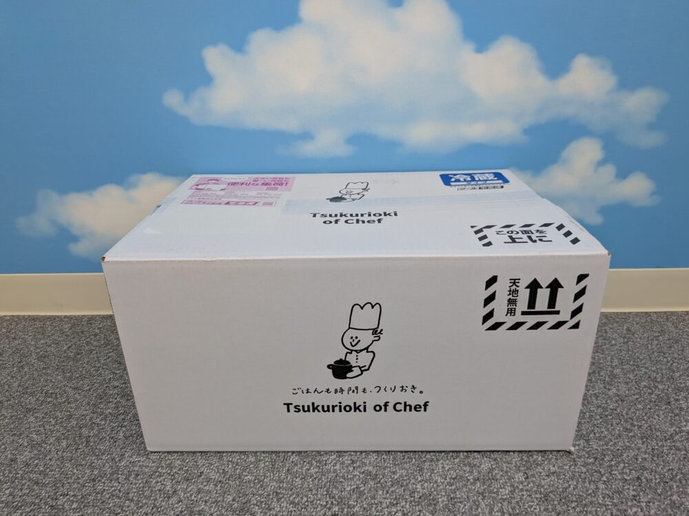 自宅に届いた「シェフの無添つくりおき」の宅配の実際の写真。珍しい白色の段ボールで、かわいいシェフのキャラクターがお鍋を持ってコンセプト「ごはんも時間もつくりおき。」という言葉と並んでいる。